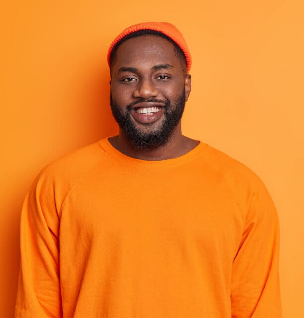 A smiling man with a beard wears an orange beanie and an orange sweatshirt, standing in front of an orange background.