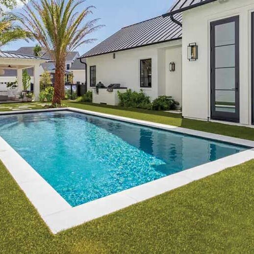A modern backyard features a rectangular swimming pool surrounded by green grass, tall palm trees, a white house with large windows, and a covered patio area in the background.