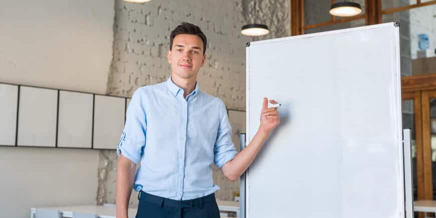 A man in a light blue shirt stands next to a blank whiteboard in a modern classroom, pointing at the board with a marker. Tables, chairs, and pendant lights are visible in the background.